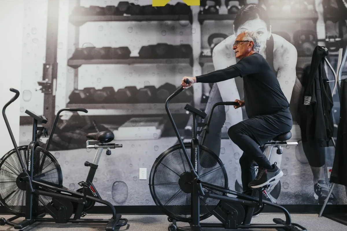 An older man pedals an air bike beside the dumbbell racks at an Alloy studio