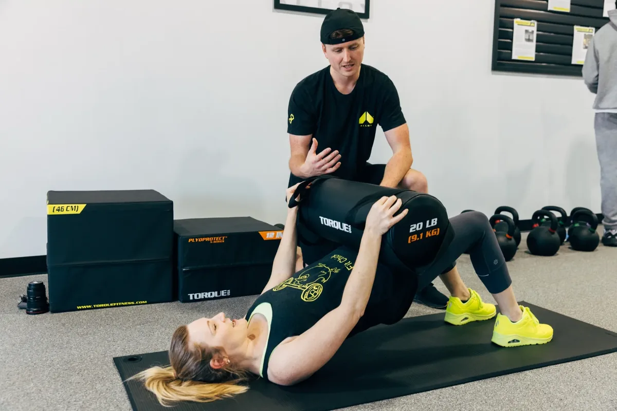 A coach kneels behind a member on a floor mat, coaching her through glute bridges with a 20 lb sandbag across her hips