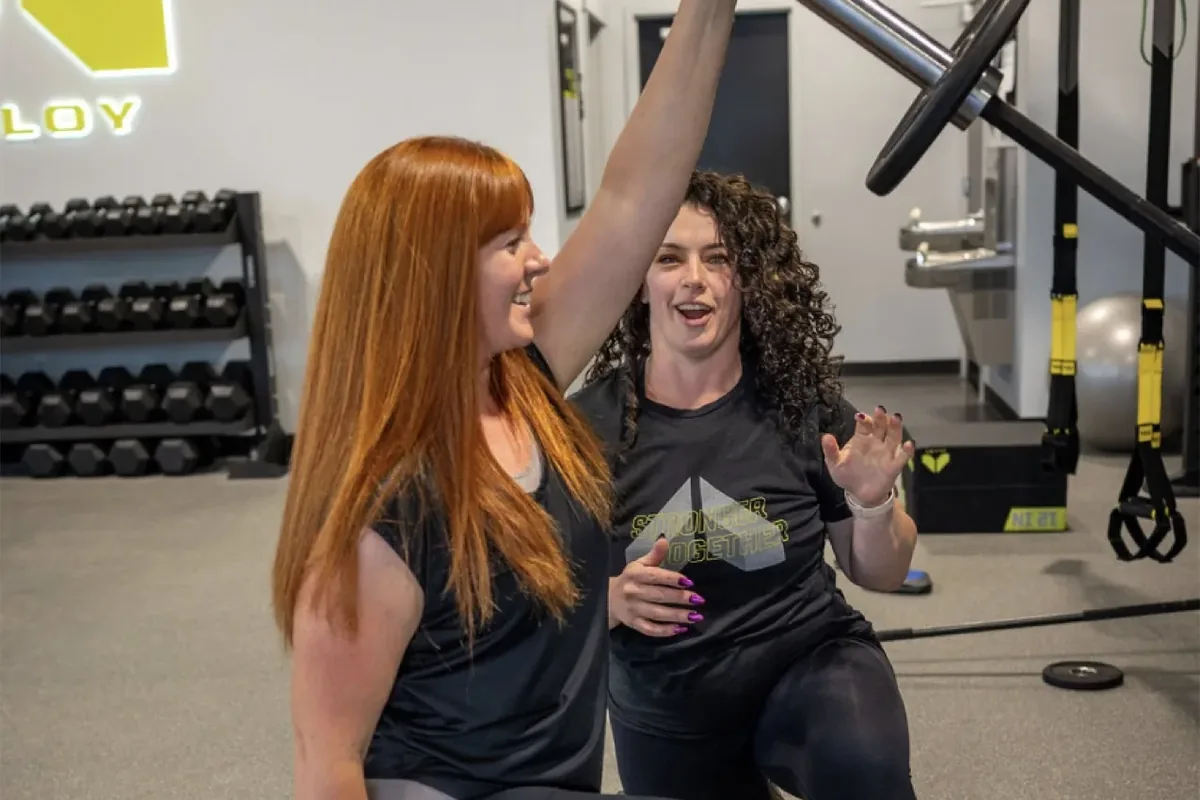 A trainer laughs with a member as she presses a bar overhead from a kneeling lunge