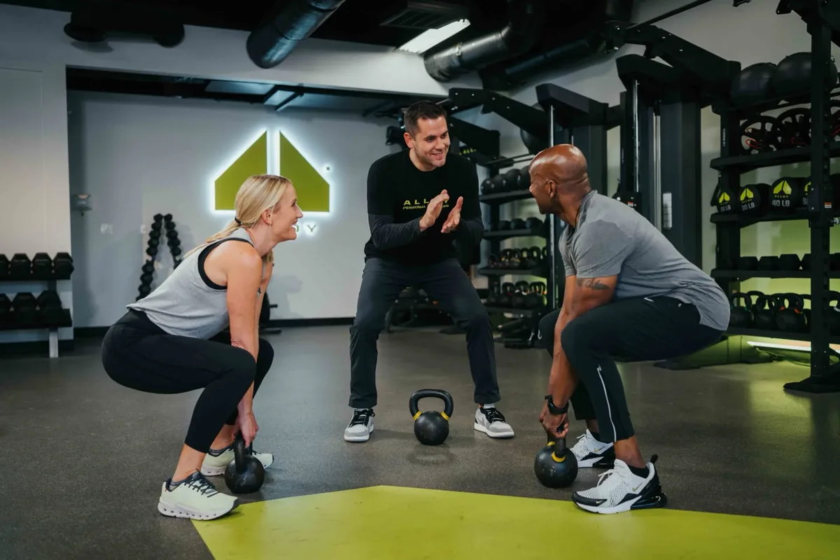 Coach encouraging two members through side-by-side kettlebell squats at Alloy Personal Training Madison East