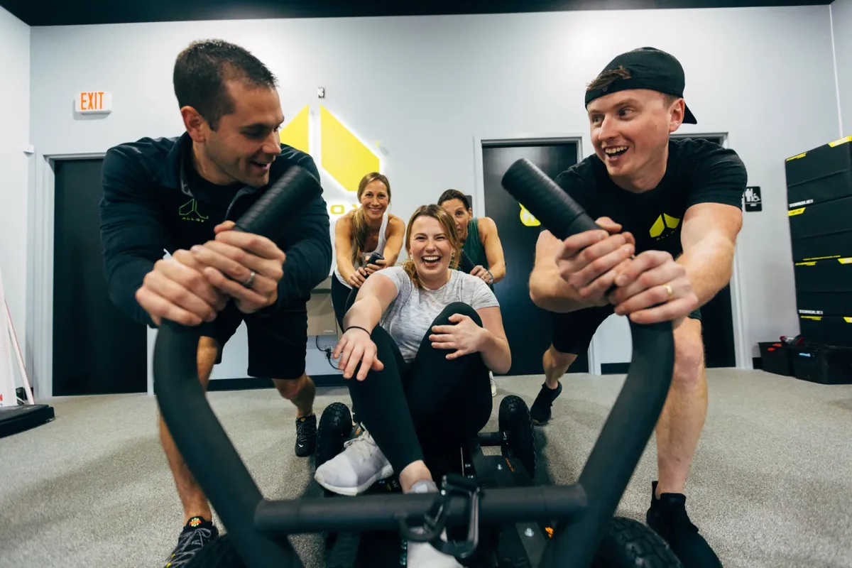 A member laughs mid sled push while her coach and training group cheer her on