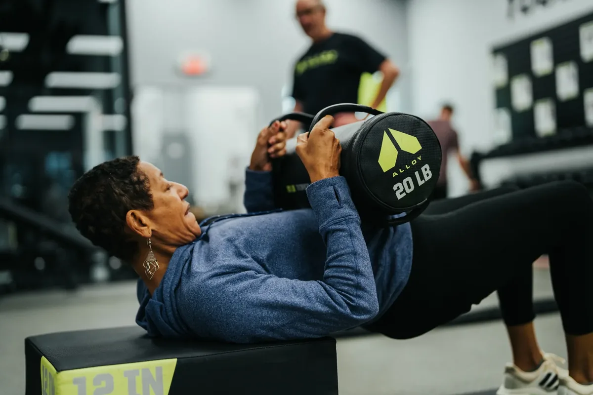 An older woman presses a 20 lb Alloy sandbag from a bench during a strength session at Alloy Personal Training Madison East.